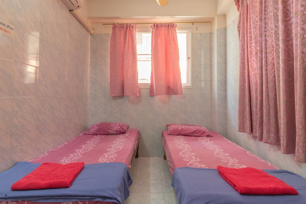 A room with two beds, each with a red towel, pink curtains, and a window with a view of the outside.