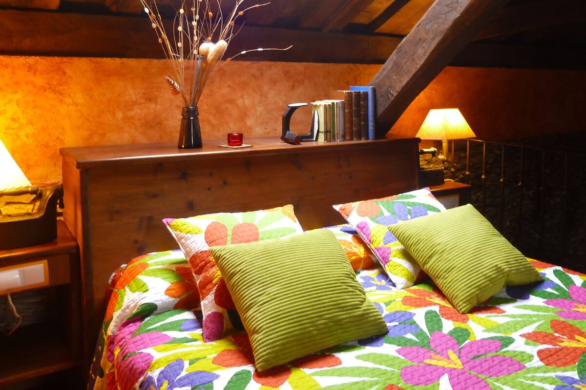 A bed with colorful floral bedding and two green pillows, a wooden headboard, a vase with branches, and a stack of books.