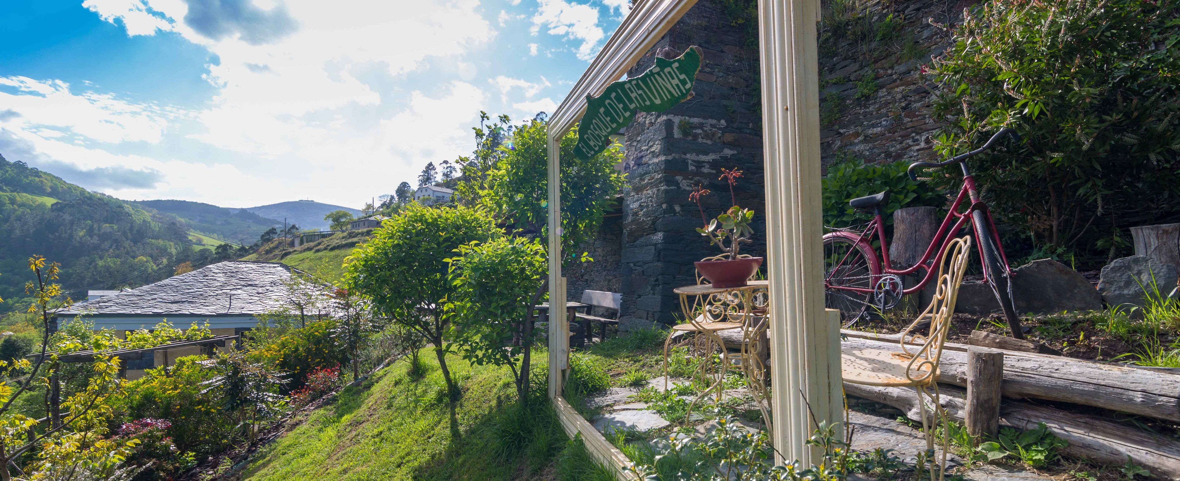 A rustic outdoor seating area with metal chairs and a table, surrounded by greenery and a stone wall.