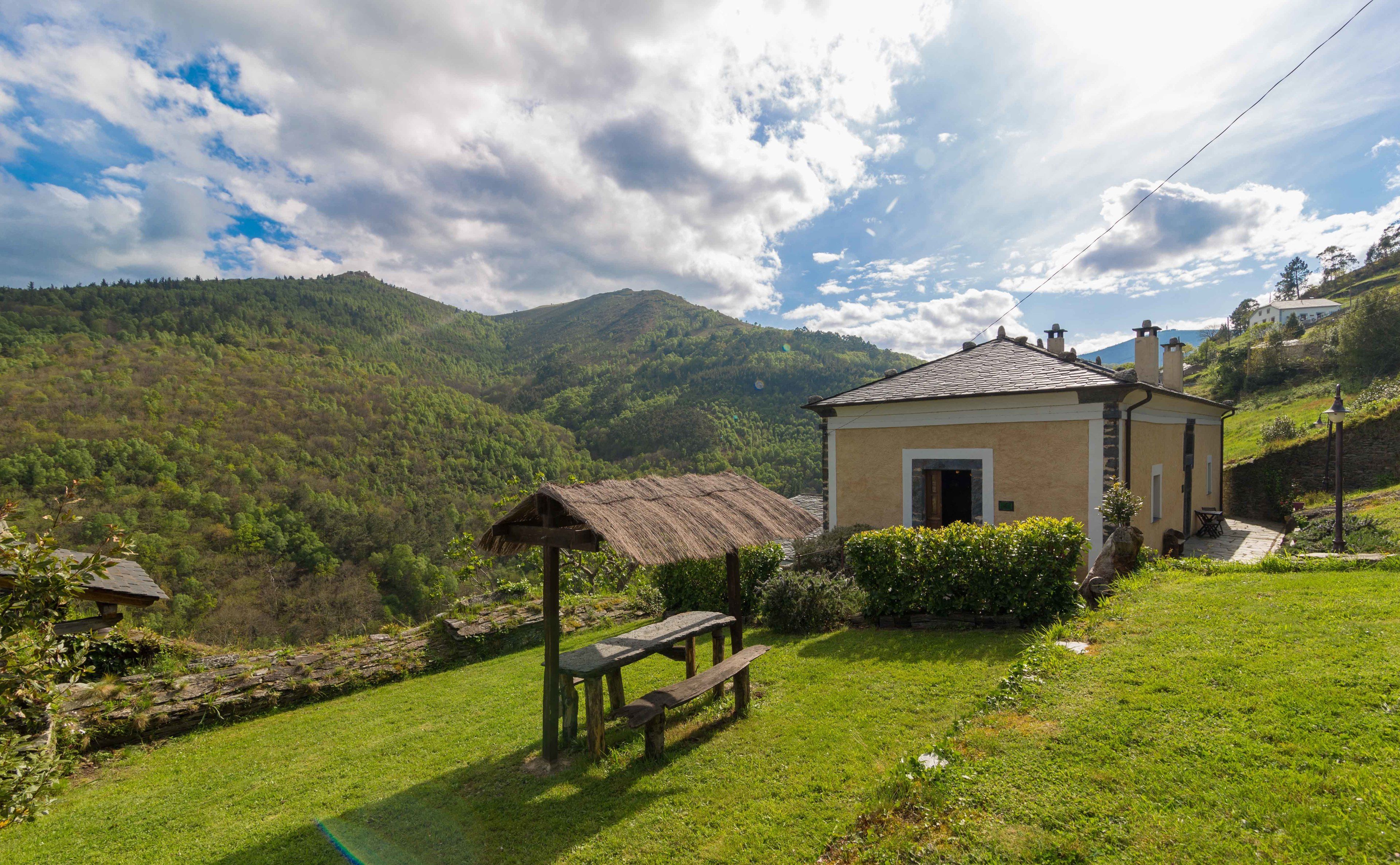 A rustic house with a thatched-roof structure and a stone bench in a lush green valley.