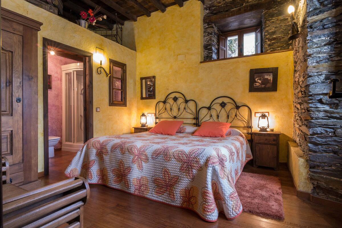 A bedroom with a large bed, wooden headboard, stone walls, and a balcony with flowers.