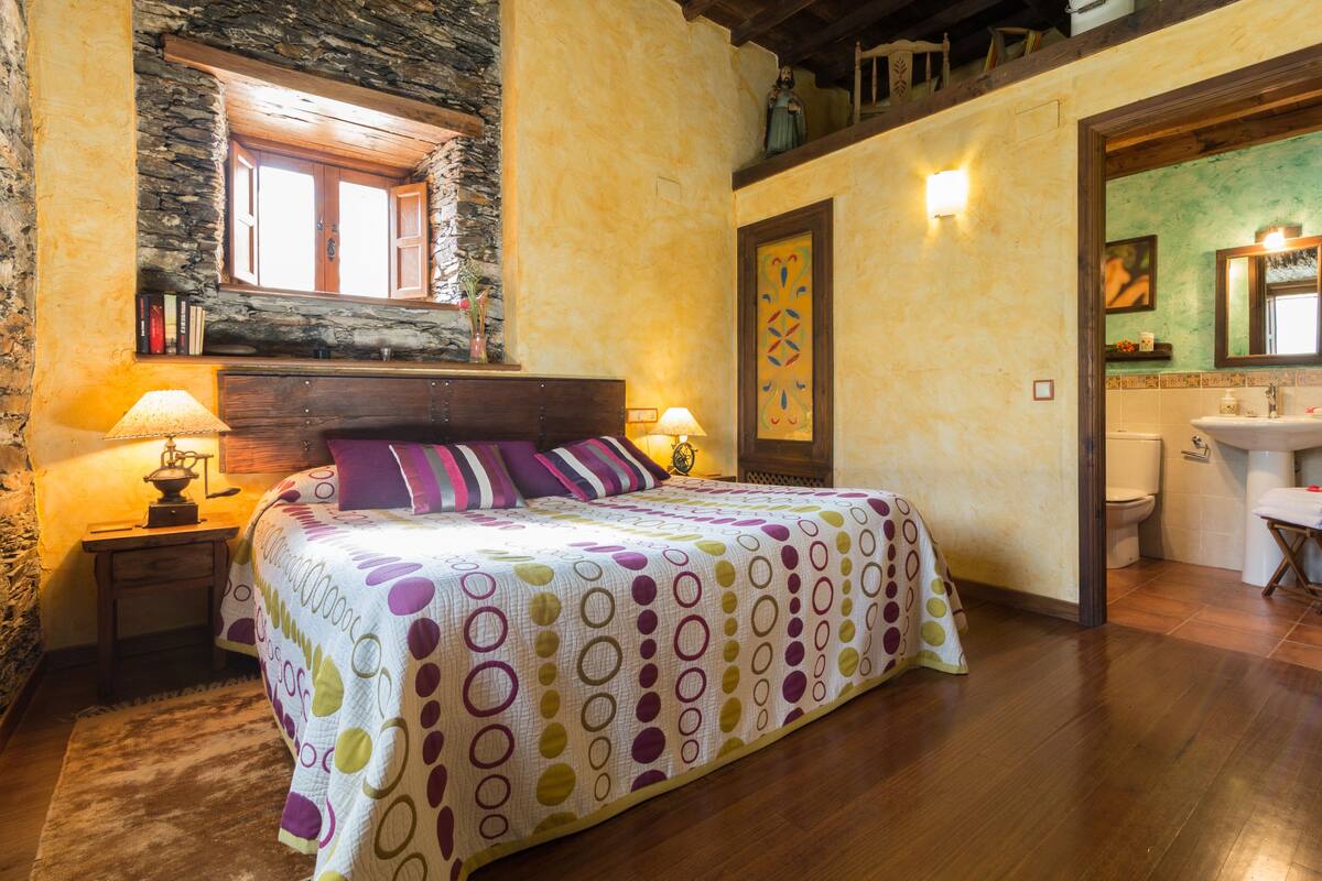 A bedroom with a bed, two bedside lamps, a stone wall, a wooden headboard, a window, a bathroom with a sink and toilet visible through an open door.