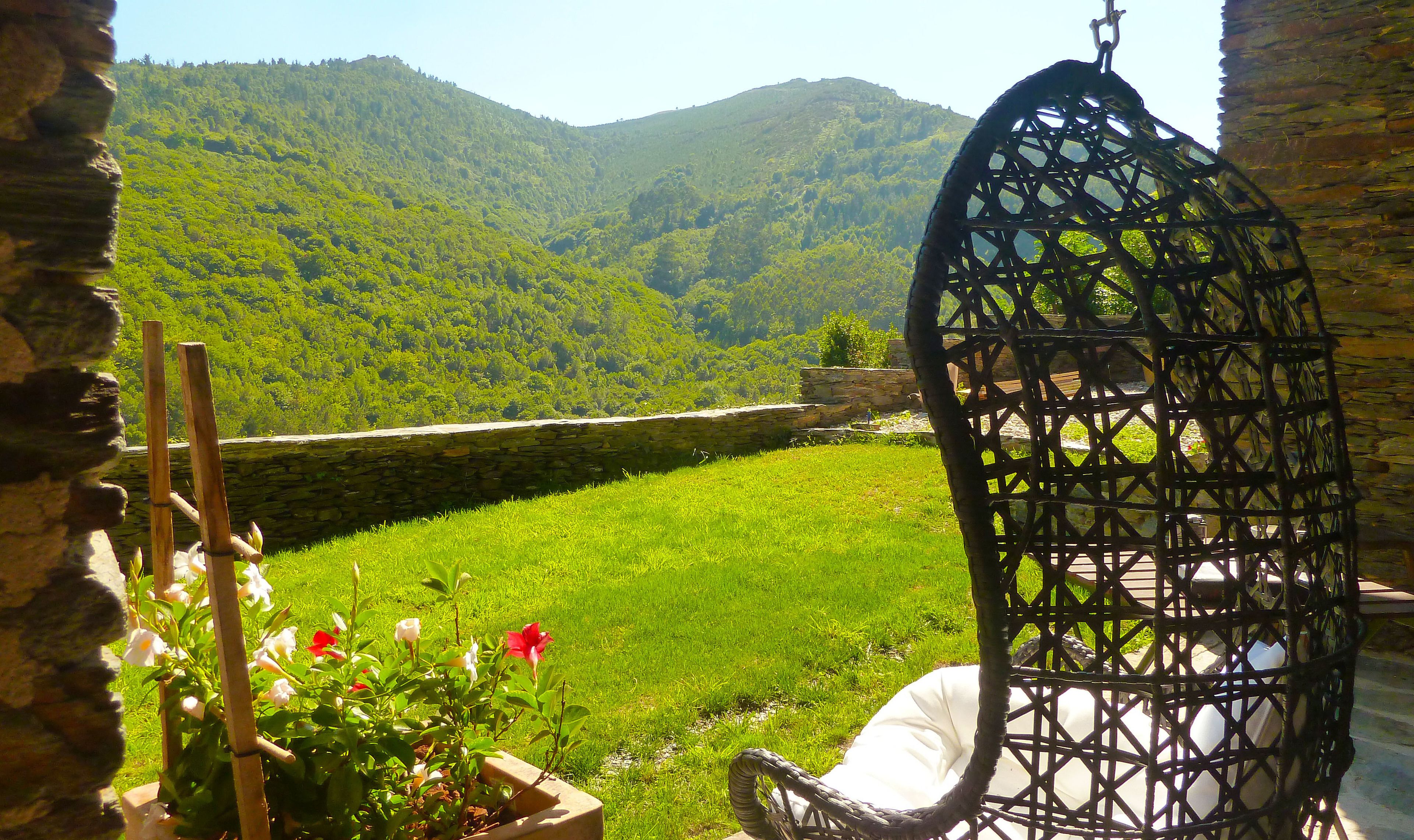 A garden with a stone wall, a hanging chair, and a view of a green valley.