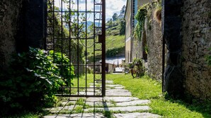 A stone pathway leading to a gated entrance with a bell above it, surrounded by greenery and a view of a hillside with buildings. - El Bosque de las Viñas (Boal)