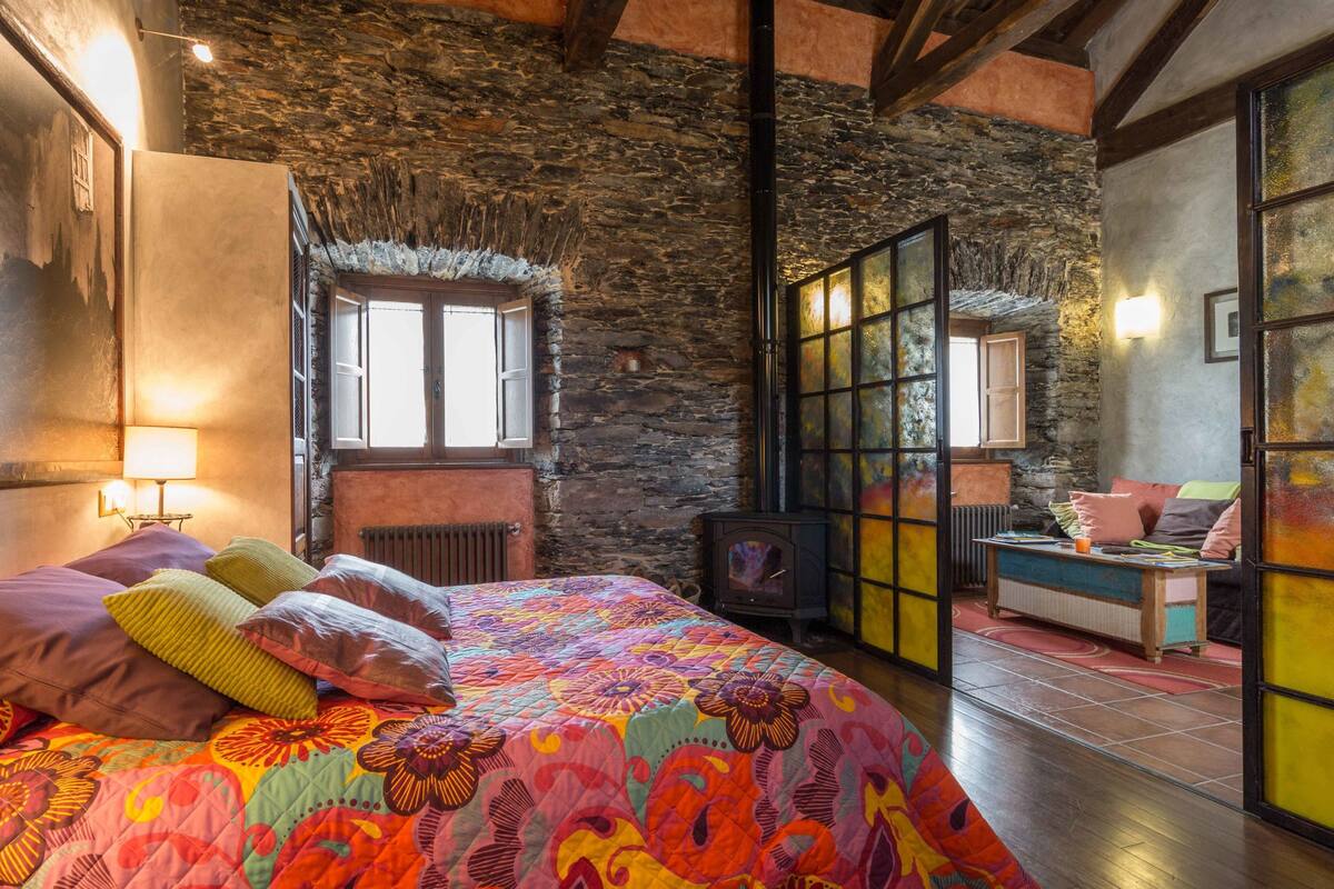 A room with a stone ceiling, a bed with colorful bedding, a window with shutters, and a view of another room through a glass door.