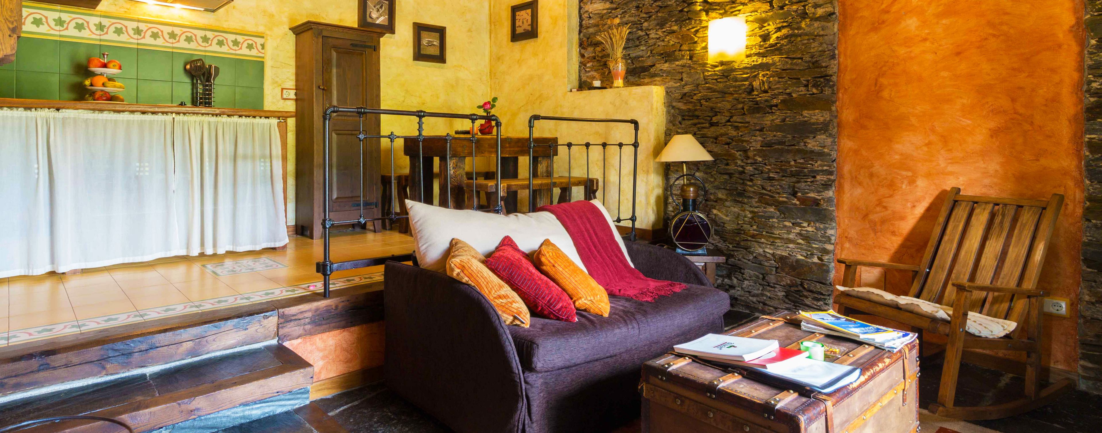 A cozy room with a sofa, a wooden rocking chair, a table with books, a lamp, and a stone wall.