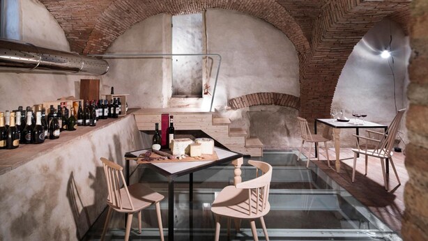 Una cava con arcos de ladrillo, una estantería para vinos larga y una mesa con queso y botellas de vino.