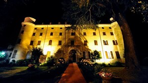 Un edificio grande e iluminado con una entrada central, rodeado de un jardín cuidado y un sendero rojo que conduce a la puerta.