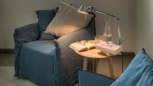 Una habitación acogedora con un sillón azul, una mesita con una lámpara y una ventana con vistas a la vegetación.
