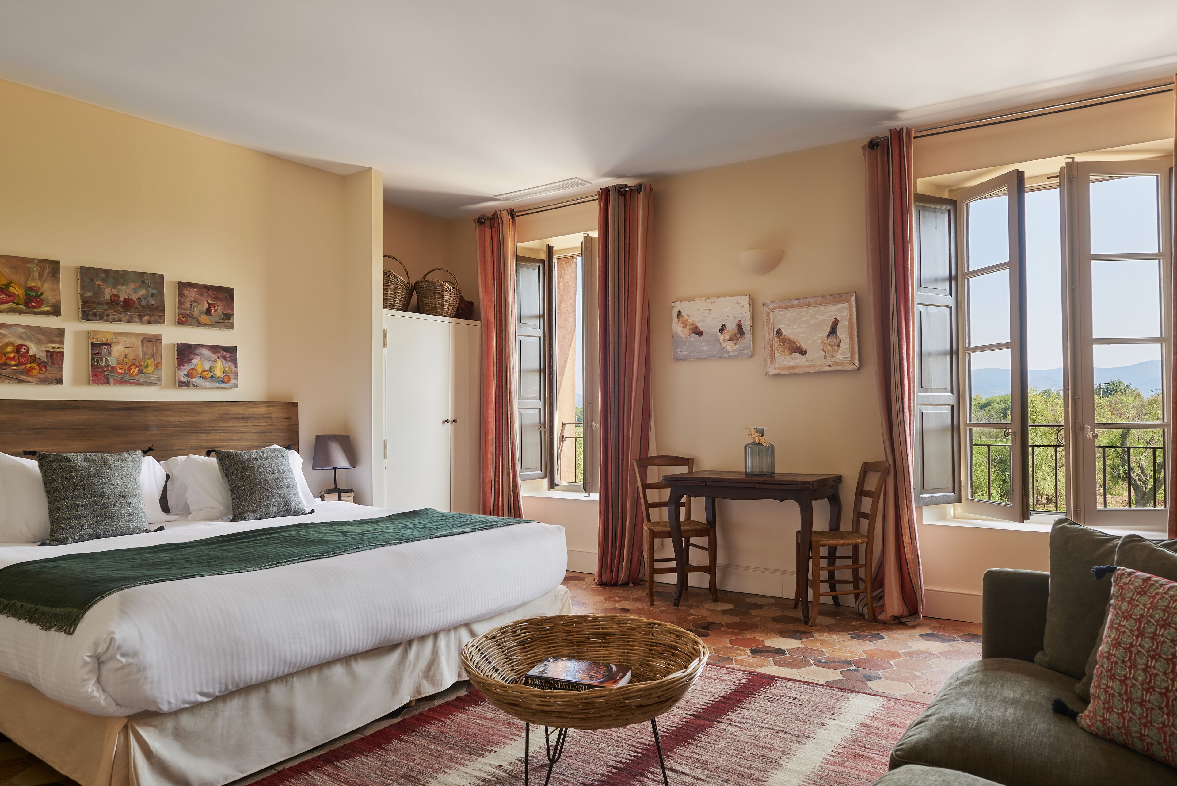 Quarto com cama, mesa de jantar, cadeiras, uma poltrona de vime e obras de arte nas paredes.