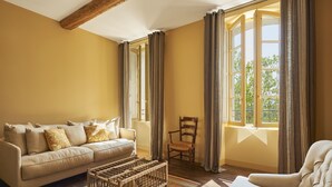 Une pièce avec un canapé beige, une table basse en bois, une chaise en bois et une grande fenêtre avec des rideaux.