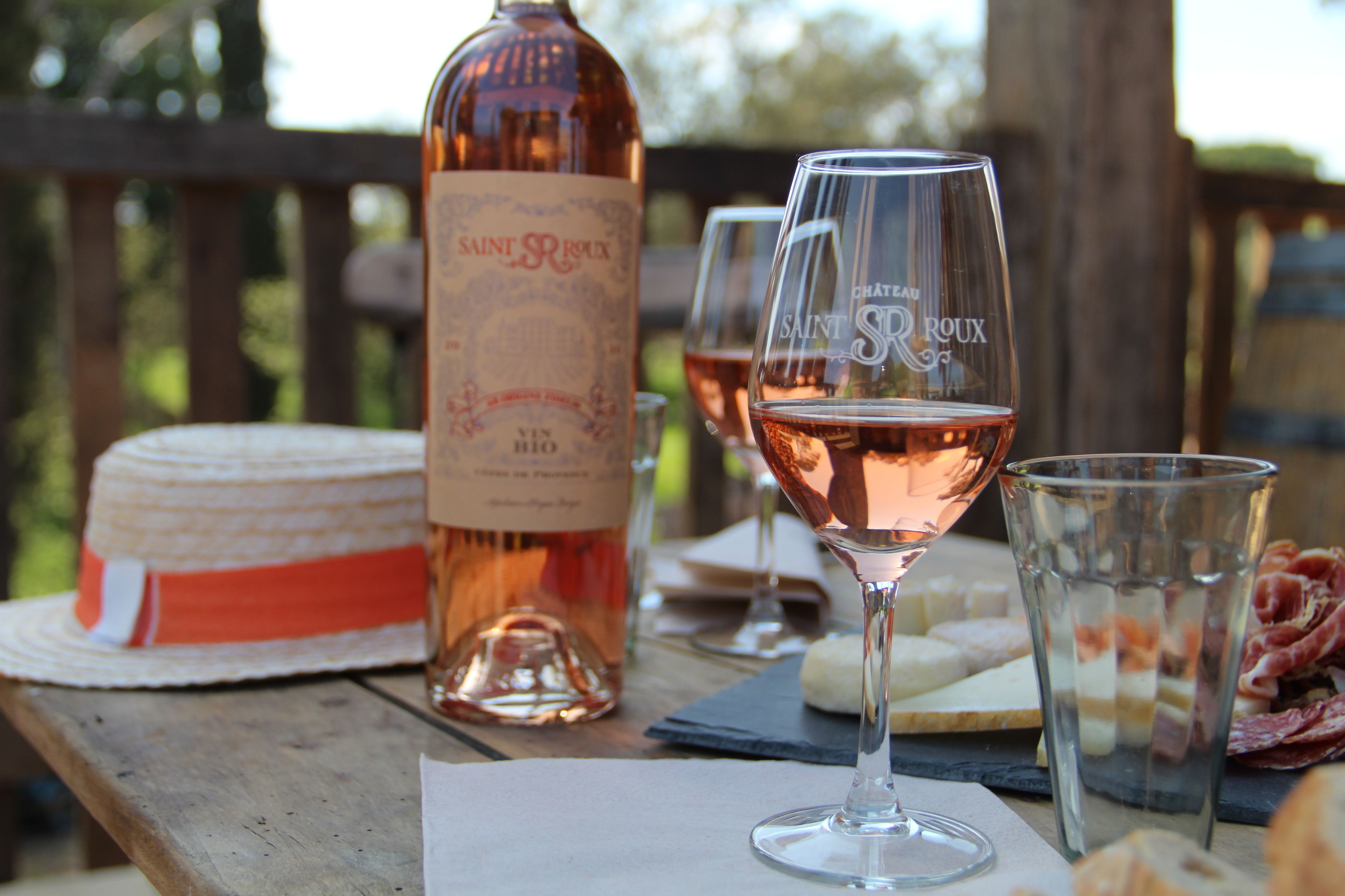 A wooden table with a bottle of Saint Roulx wine, two glasses of rosé, a hat, and a cheese board with various cheeses and cured meats.