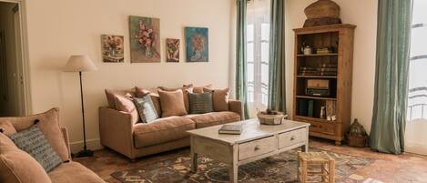 Un salón con un sofá beige, una mesa de centro y un tapete estampado. Hay cuadros en la pared y un aparador de madera con libros y adornos.