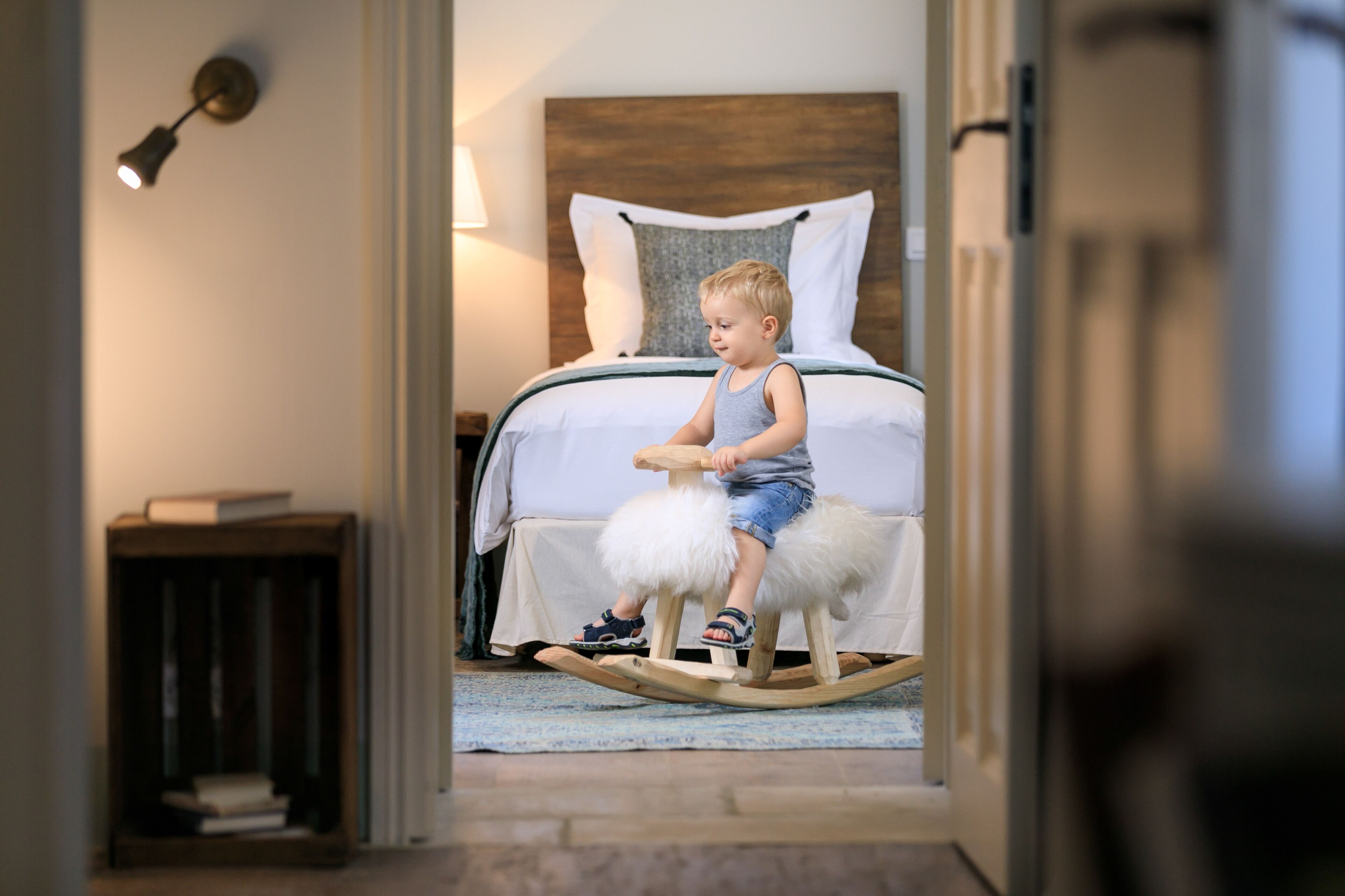 A child on a rocking horse in a bedroom with a wooden headboard, a bedside table, and a lamp.