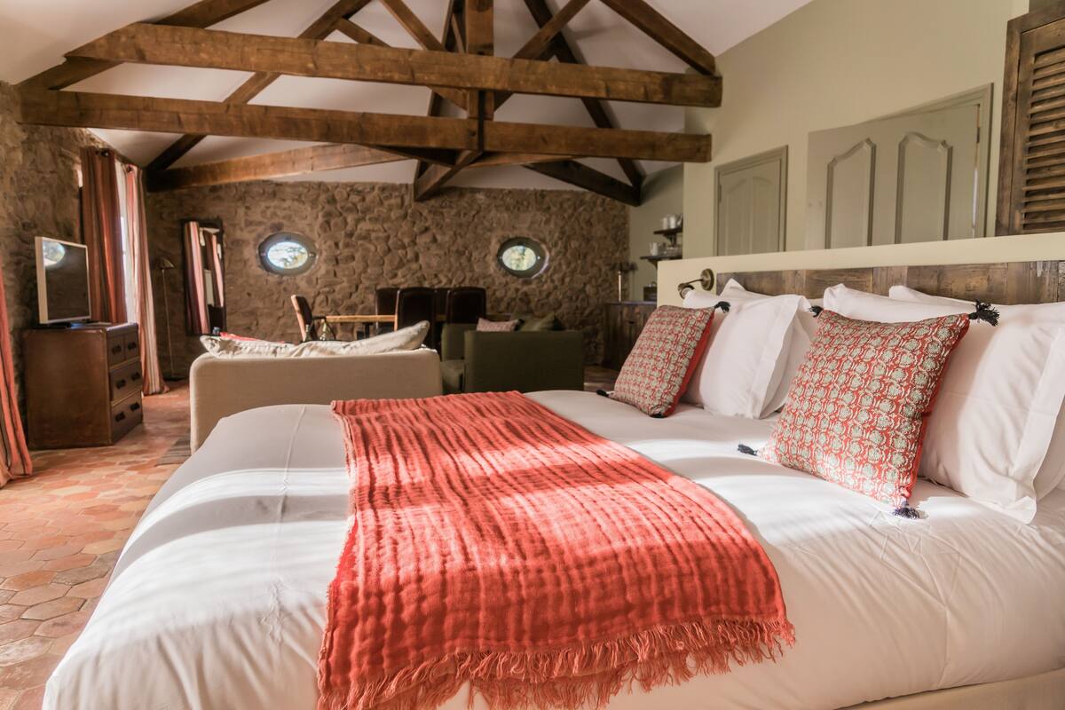 Un dormitorio con una cama grande, una pared de piedra, vigas de madera, un televisor y una pequeña mesa con sillas.