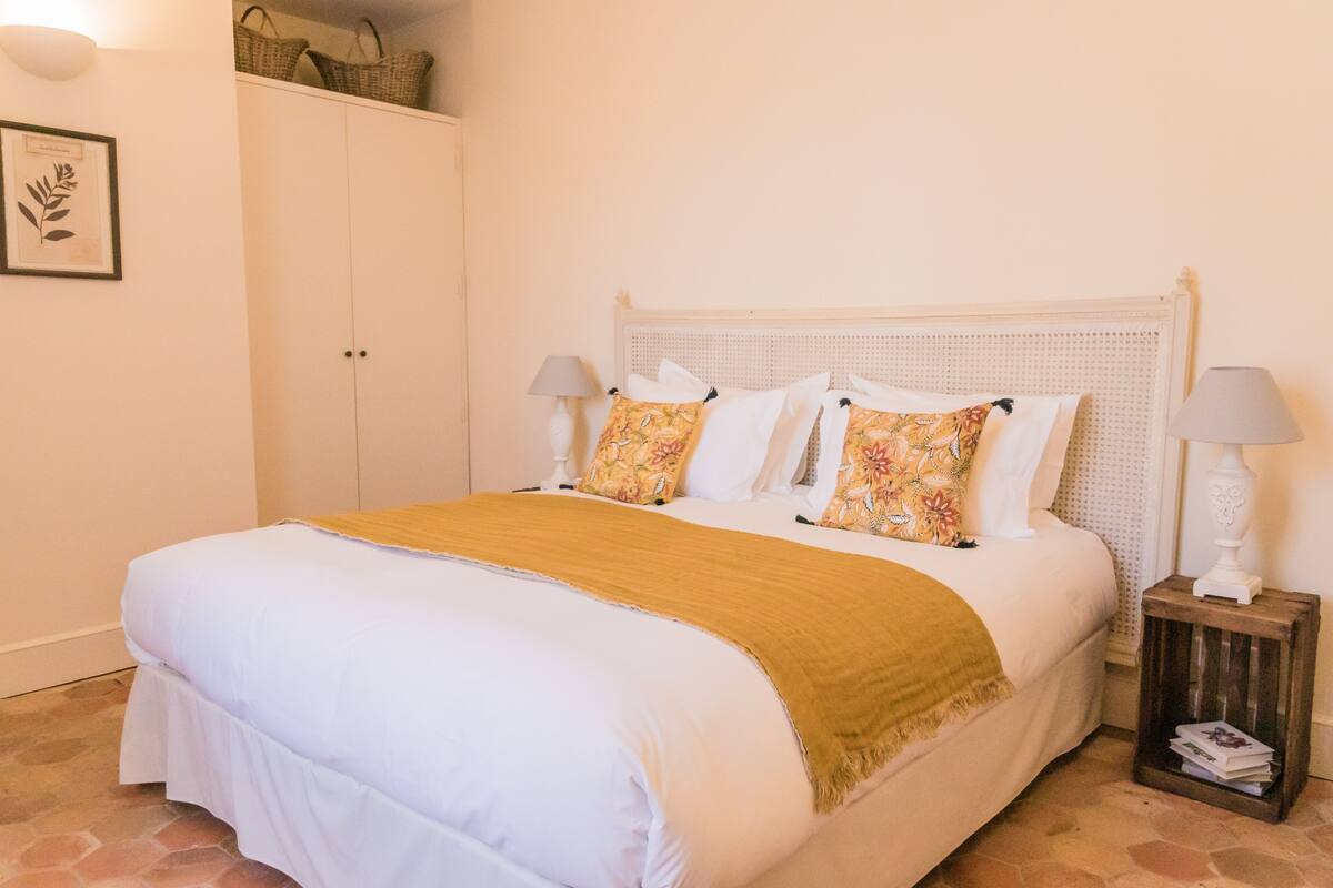 Una cama bien hecha con colcha blanca y color mostaza, dos almohadas a juego y un cabecero de madera. Hay dos lámparas de noche a cada lado de la cama.
