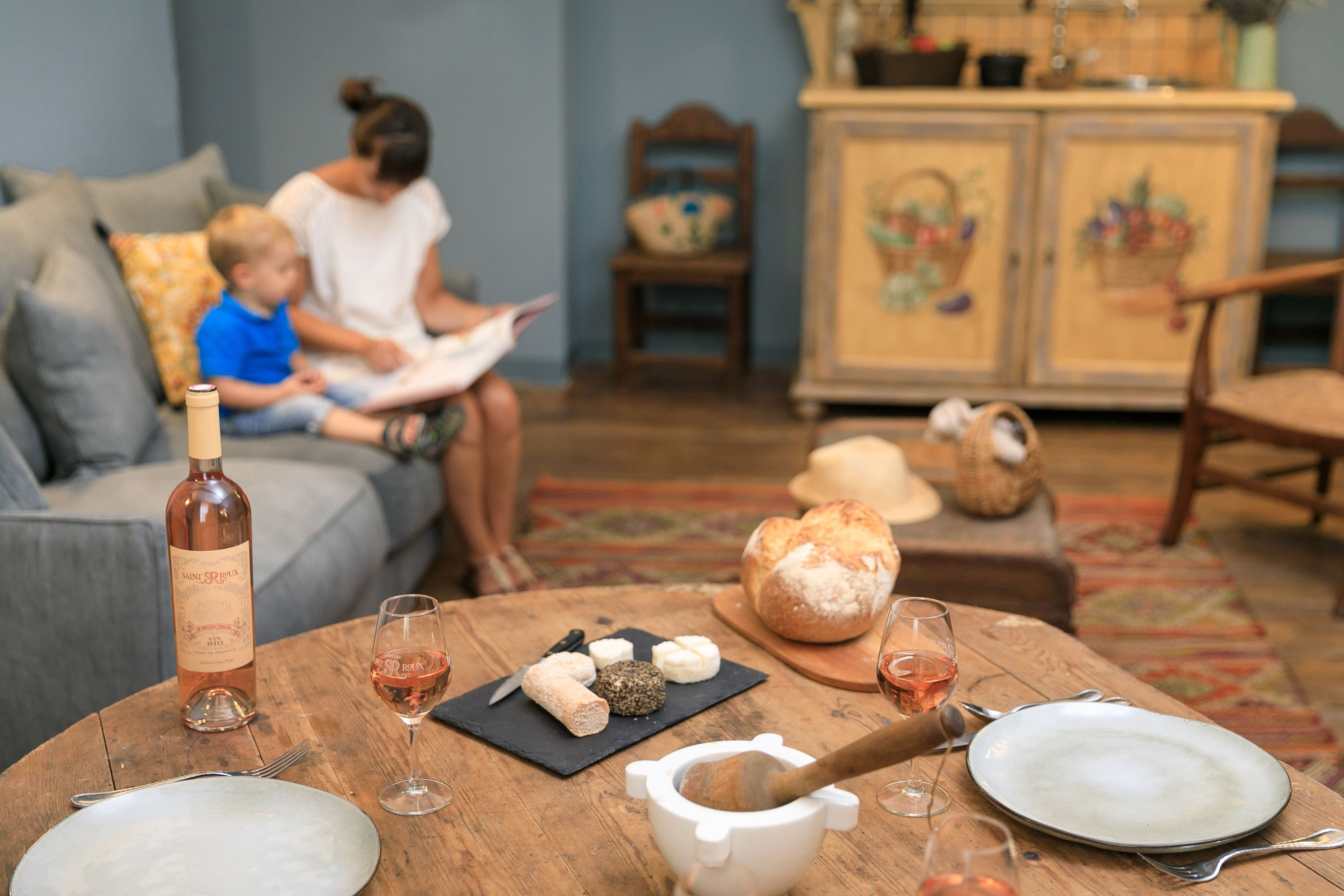 Un salón acogedor con una mesa de madera dispuesta para una comida, que incluye una botella de vino, queso, pan y un ambiente rústico.