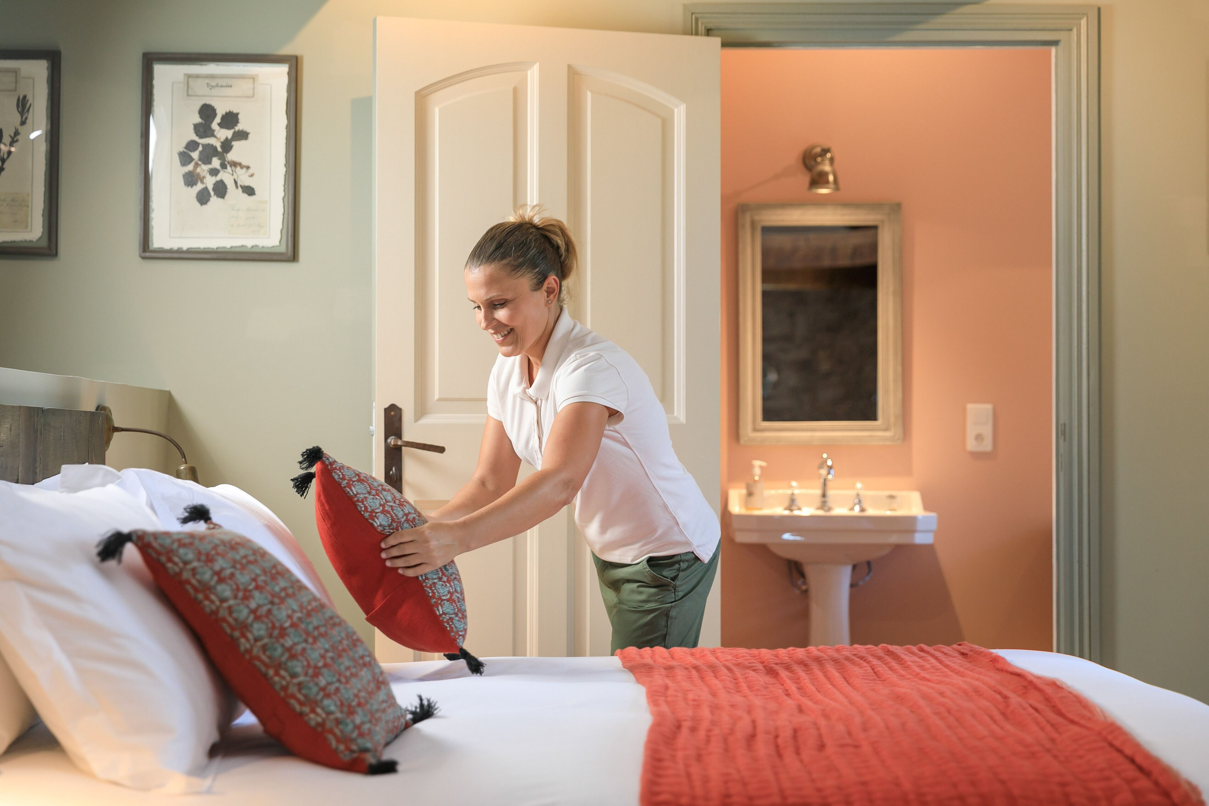 A hotel room with a bed, pillows, a red blanket, a sink, and framed botanical prints on the wall.