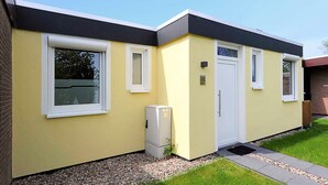 A modern single-story building with a flat roof, a yellow facade, and a black trim. There are two windows on the front, a door, and a small utility box to the left. - Vacation home Nicole - Holiday homes Friesensterne (Esens)