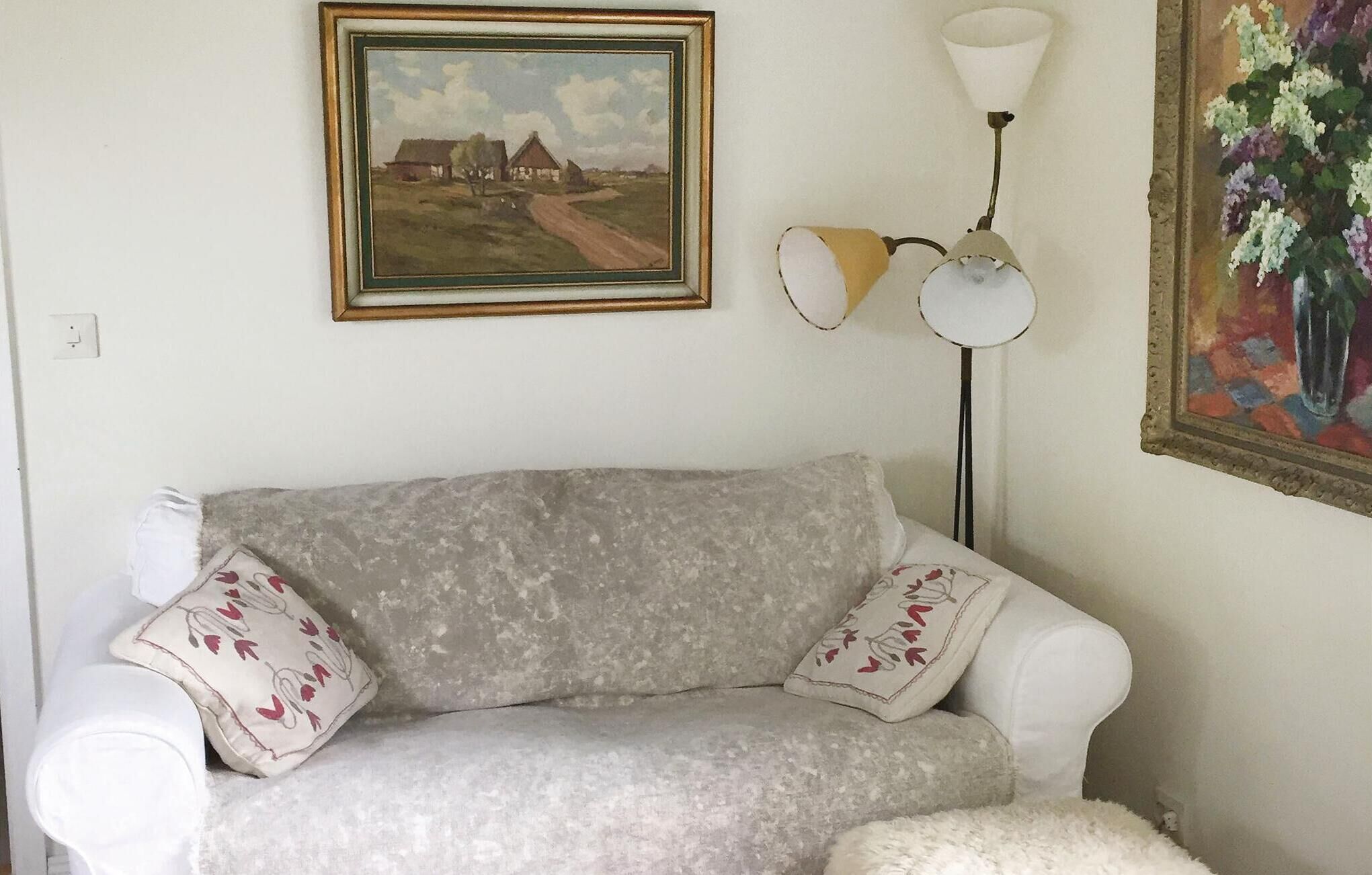 A white sofa with floral pillows, a framed landscape painting, a floor lamp, and a floral painting on the wall.