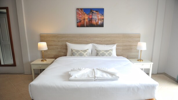 A neatly made bed with white linens and a folded towel, two bedside tables with lamps, a wooden headboard, and a cityscape painting on the wall. - Drop Inn Bangkok (Bangkok)