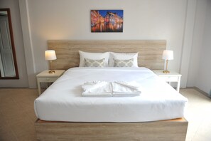 A neatly made bed with white linens and a folded towel, two bedside tables with lamps, a wooden headboard, and a cityscape painting on the wall. - Drop Inn Bangkok (Bangkok)