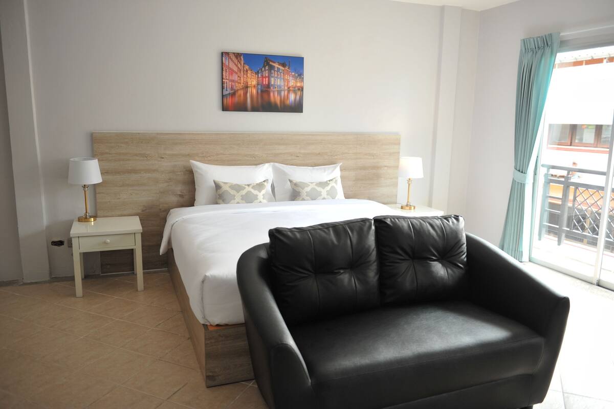 A hotel room with a bed, a black leather chair, a nightstand with a lamp, and a painting on the wall.