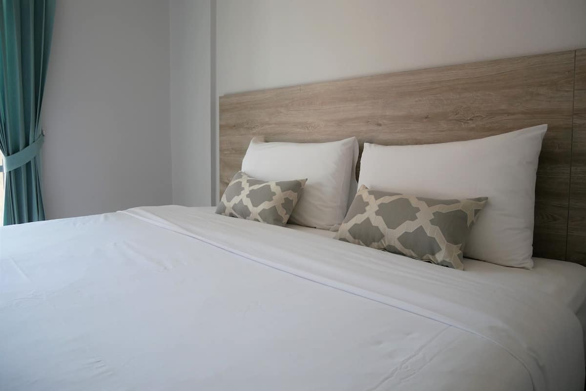 A neatly made bed with white bedding and two patterned pillows, a wooden headboard, and a green curtain partially visible.
