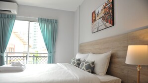 A hotel room with a bed, a nightstand, a lamp, a painting on the wall, and a window with curtains. - Drop Inn Bangkok (Bangkok)