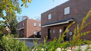 A modern brick building with a flat roof and a black door. There are windows on the second floor and a small sign on the door. - Holiday house Nordseewoge - Holiday homes North Sea Trio (Esens)