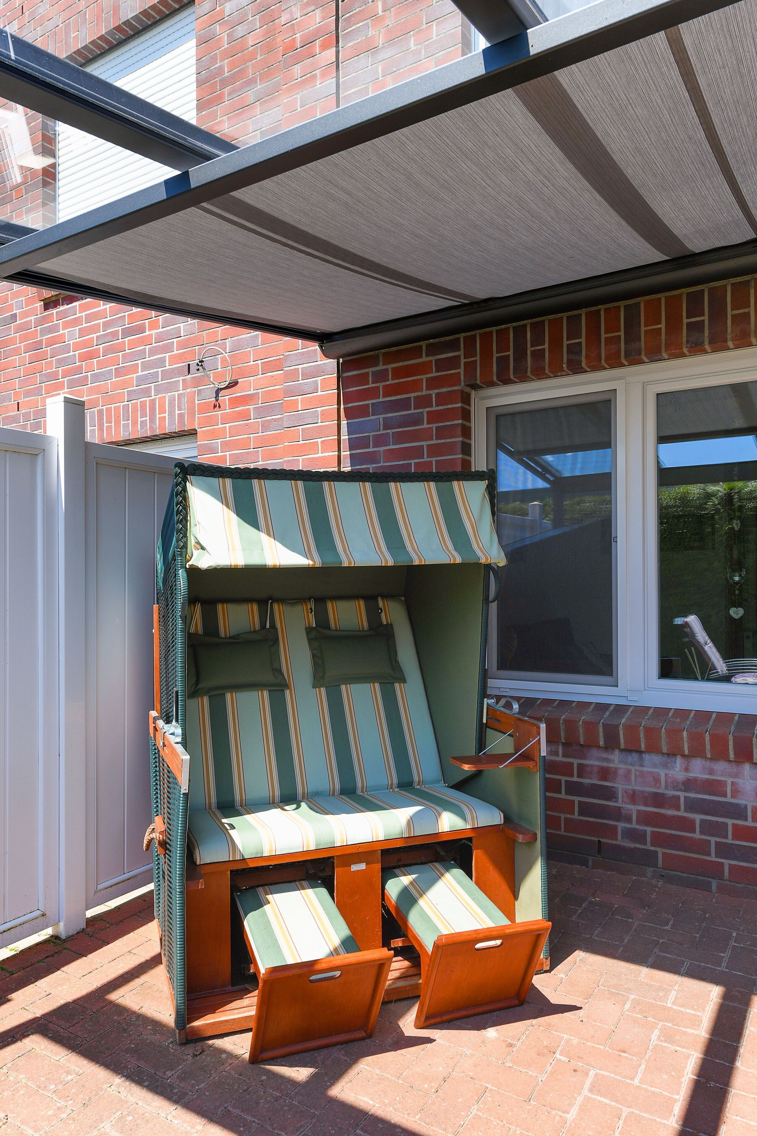 Een zonnig terras met een gestreepte lounge fauteuil en voetenbankje, een overdekte patio en een bakstenen gebouw op de achtergrond.