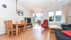 A modern living room with a wooden dining table, chairs, a red armchair, a black sofa, and a TV. - Holiday house Nordseewoge - Holiday homes North Sea Trio (Esens)