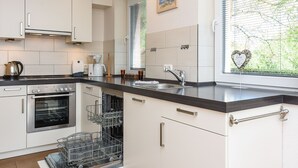 A modern kitchen with white cabinets, a stainless steel oven, a dishwasher, and a window with blinds. - Holiday house Nordseewoge - Holiday homes North Sea Trio (Esens)