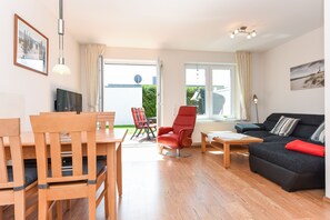 Un salon moderne avec un ensemble de salle à manger en bois, une chaise rouge, un canapé noir et une vue sur l’extérieur, accessible par une porte vitrée.
