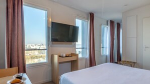 Habitación de hotel con cama, escritorio con televisor, ventana con cortinas y vista a la ciudad.