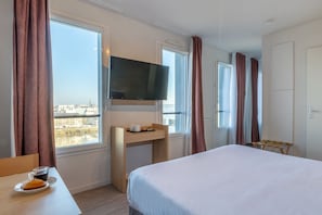 Habitación de hotel con cama, escritorio con televisor, ventana con cortinas y vista a la ciudad.