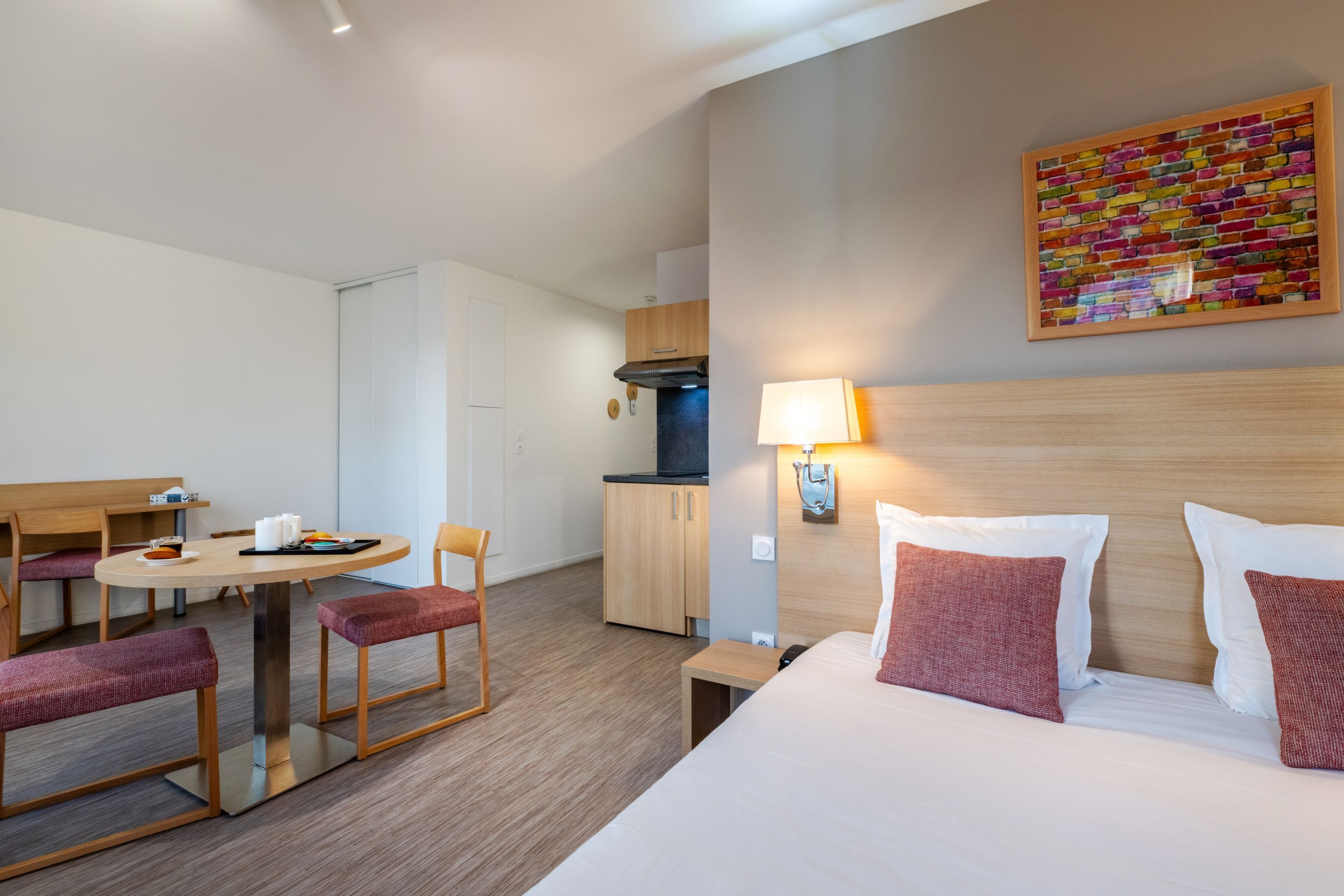 Een hotelkamer met een bed, bureau, stoel, klein tafeltje, lamp en een kitchenette.