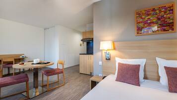 A hotel room with a bed, a desk, a chair, a small table, a lamp, and a kitchen area.