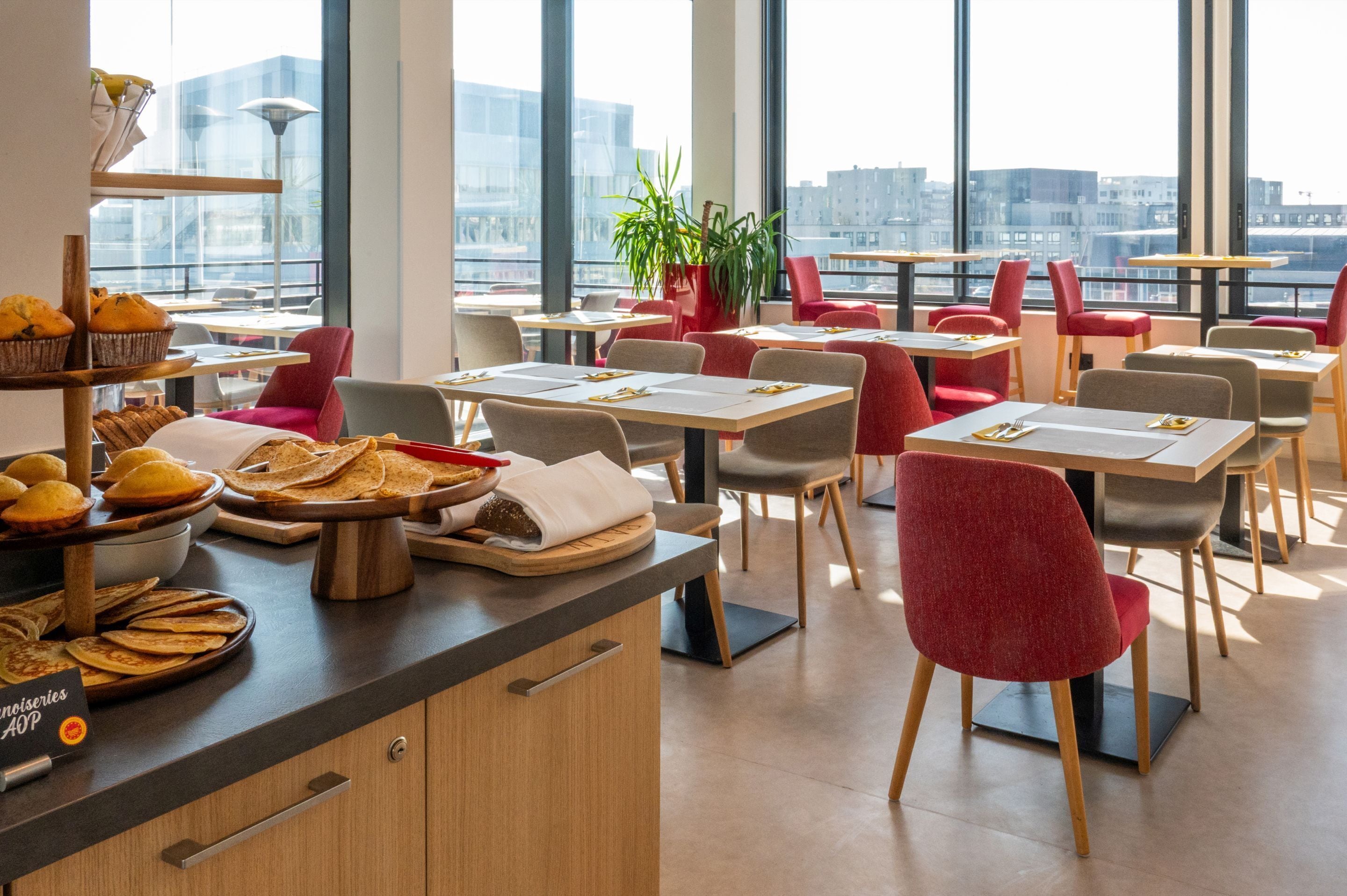 A modern restaurant with high tables and chairs, a variety of food displayed on the counter, and large windows offering a city view.