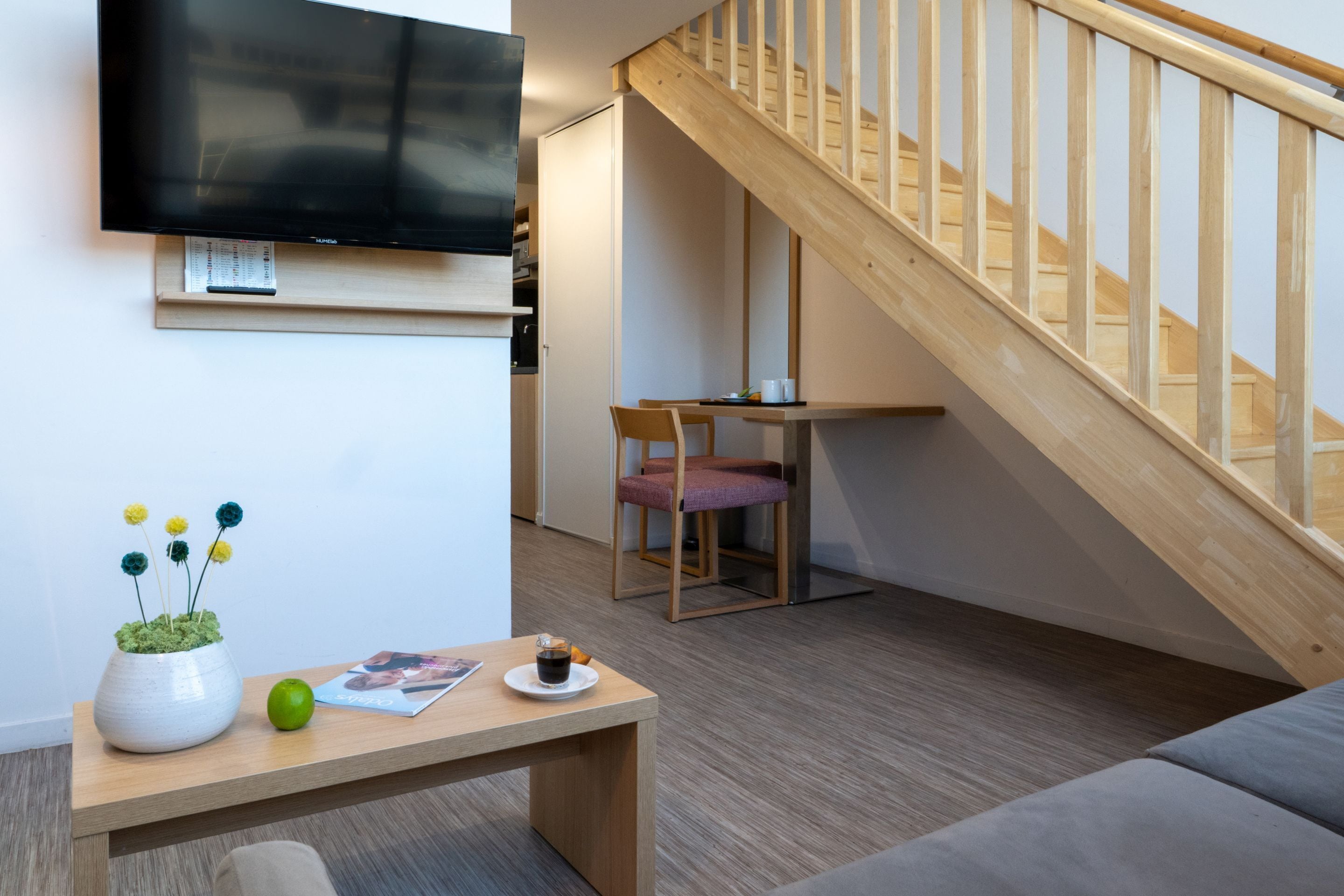 Ein modernes Wohnzimmer mit einer Holztreppe, einem an der Wand befestigten Flachbildfernseher, einem kleinen Tisch mit einer Topfpflanze und einer Tasse sowie einem grauen Sofa.