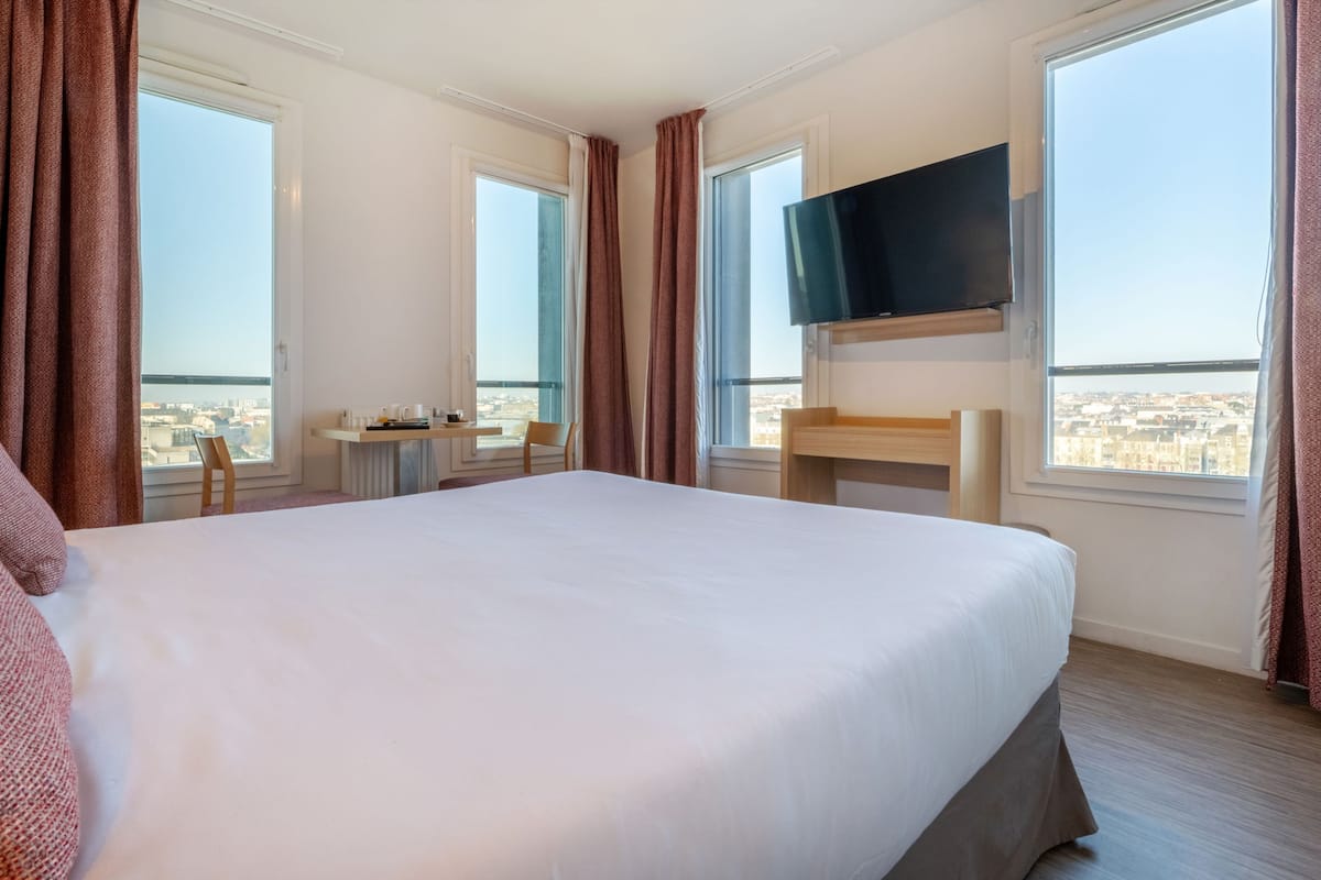 A hotel room with a large bed, a desk with a chair, a TV mounted on the wall, and large windows offering a city view.