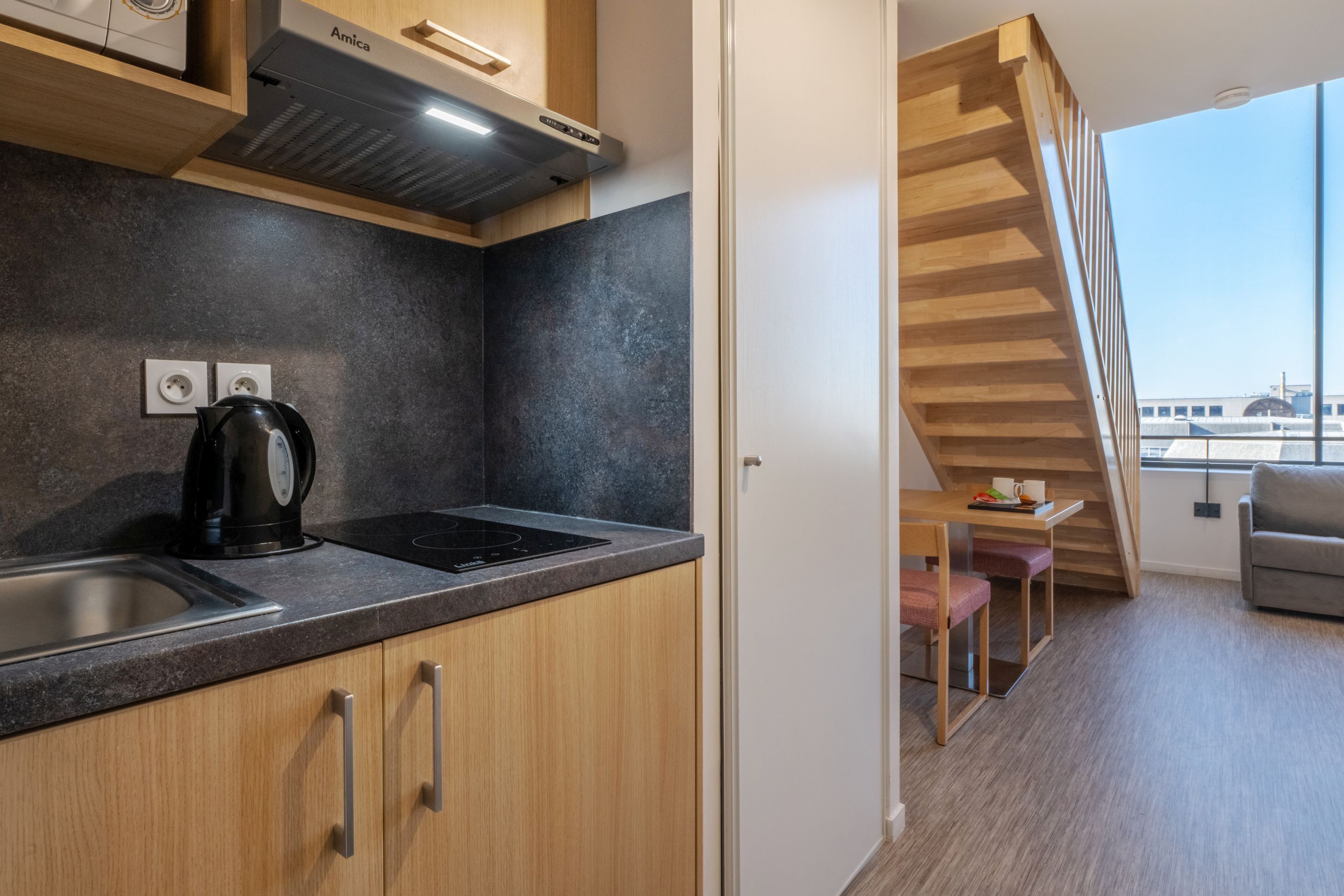 A modern kitchen with a built-in stove, a coffee maker, and wooden cabinets. There is a staircase leading to a balcony with a view of a building.