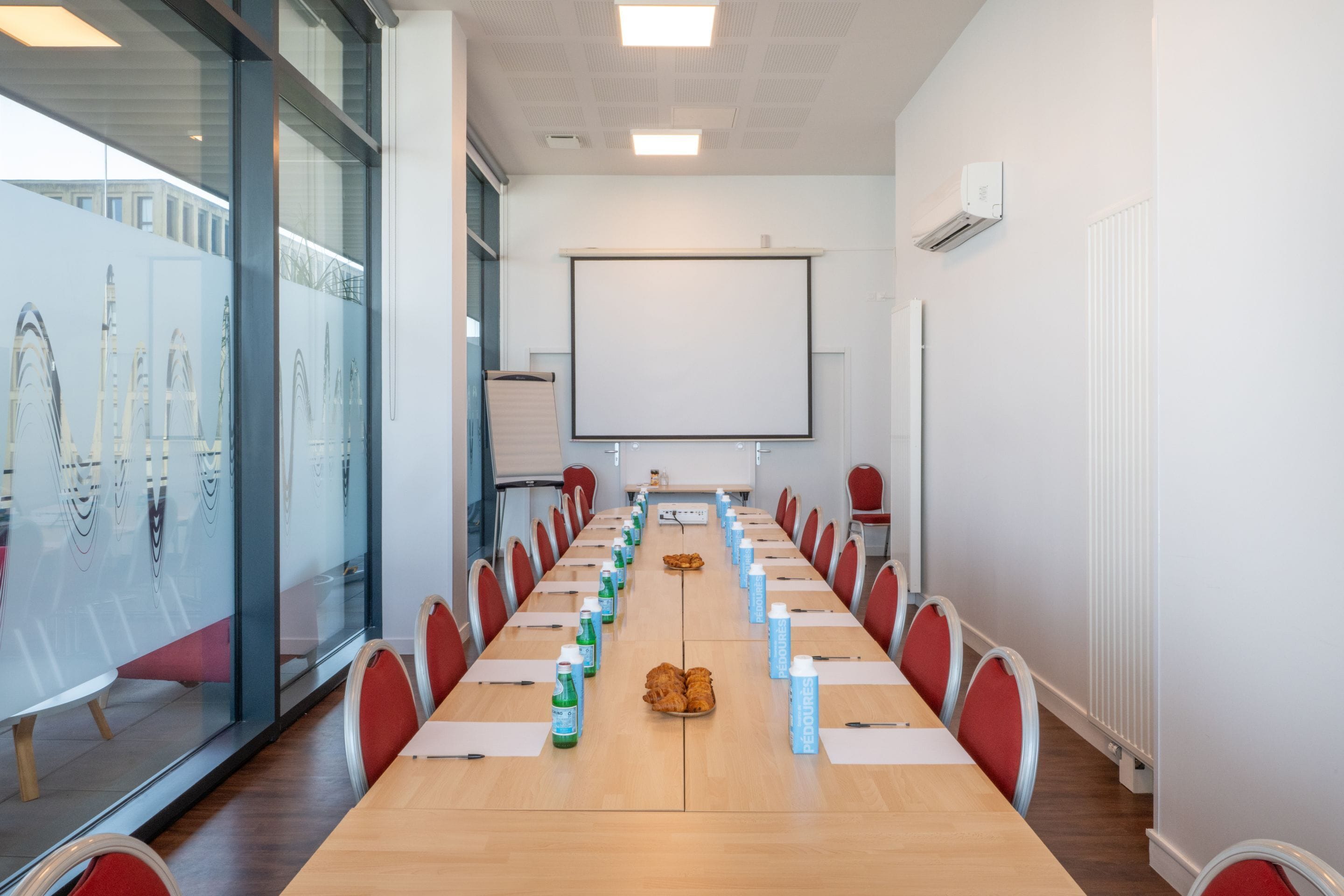 A conference room with a long table, red chairs, and a projector screen.