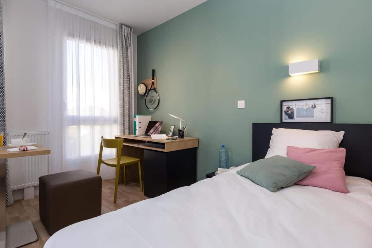 A hotel room with a bed, a desk, a chair, a radiator, and a window with curtains.