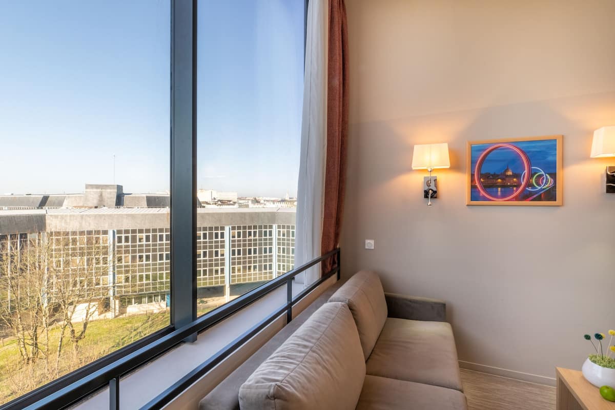 A modern hotel room with a large window, a sofa, a small table with a vase, and two wall-mounted lamps.