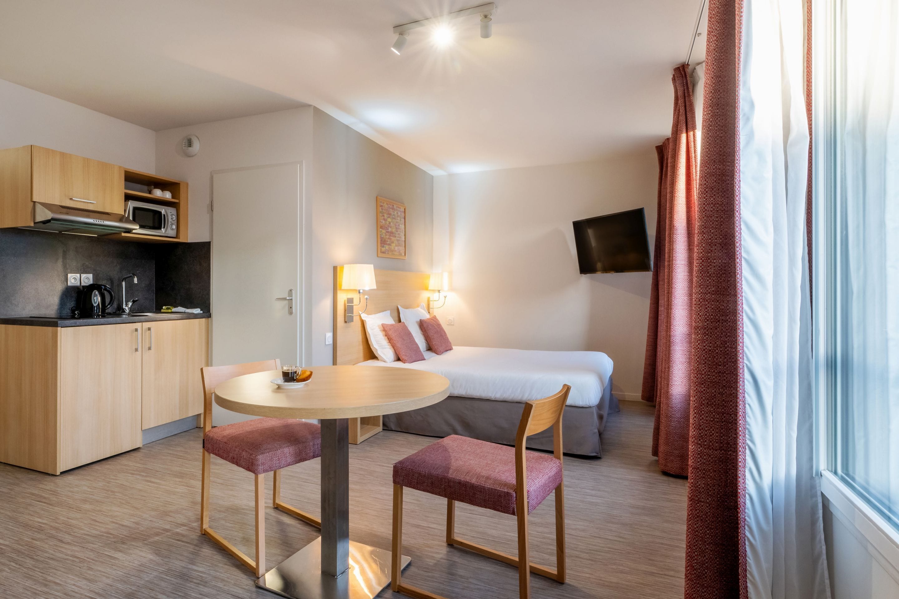 A compact hotel room with a kitchenette, a dining area with two chairs, a bed, a TV mounted on the wall, and a window with curtains.