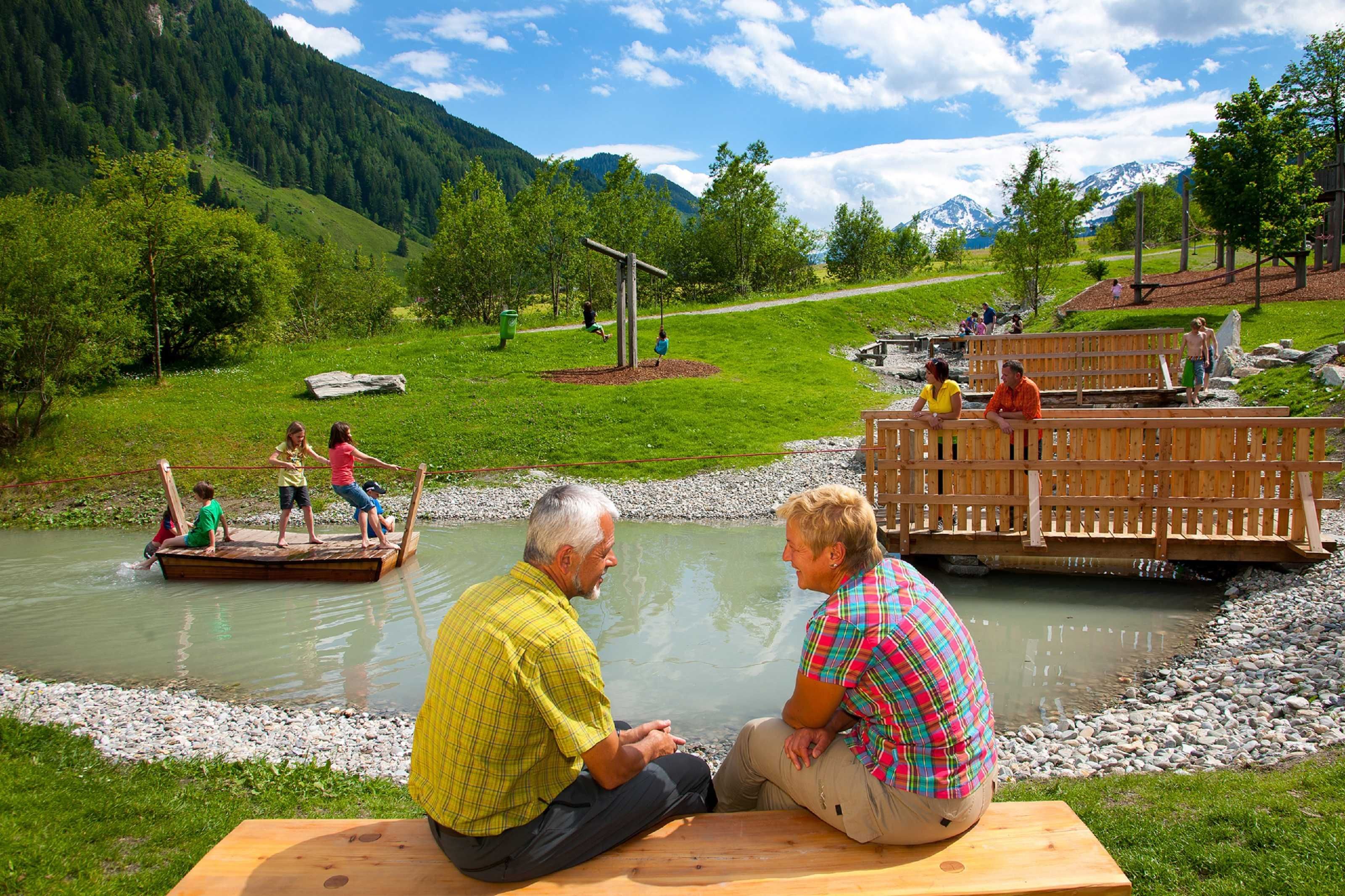 Un entorno junto al río con un puente de madera, mesas de picnic y un paisaje natural.
