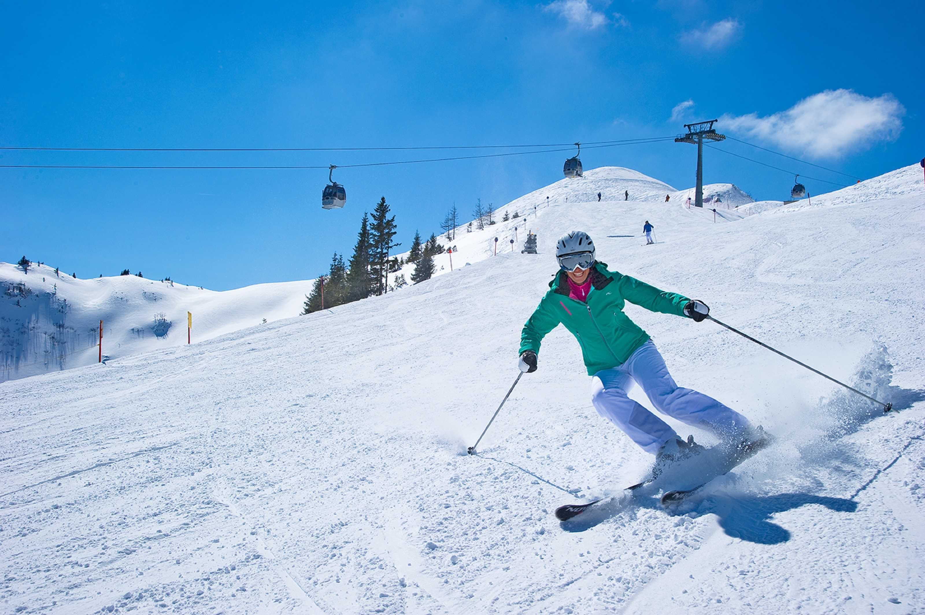 Eine Person fährt auf Skiern einen verschneiten Hang hinunter, im Hintergrund sind Skilifte zu sehen.