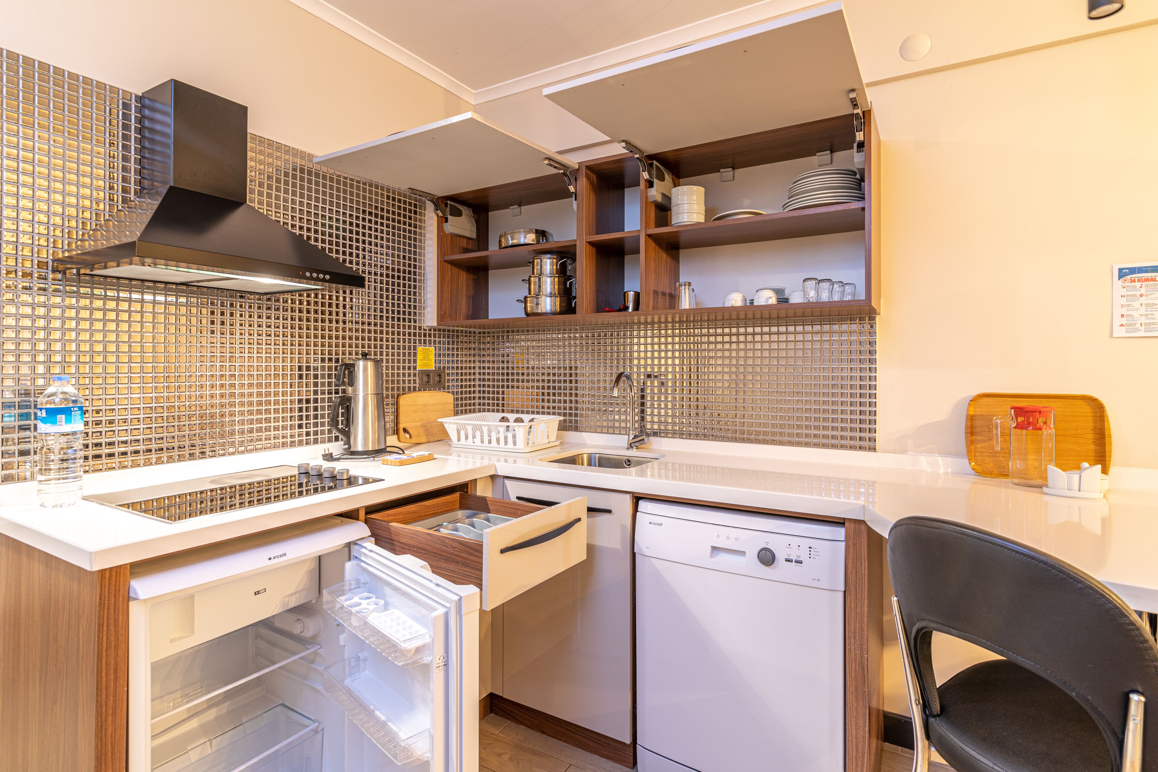 Eine kompakte Küche mit Geschirrspüler, Spüle und Dunstabzugshaube. Über der Spüle befinden sich Holzregale und ein Kühlschrank mit einer Gefriellade.