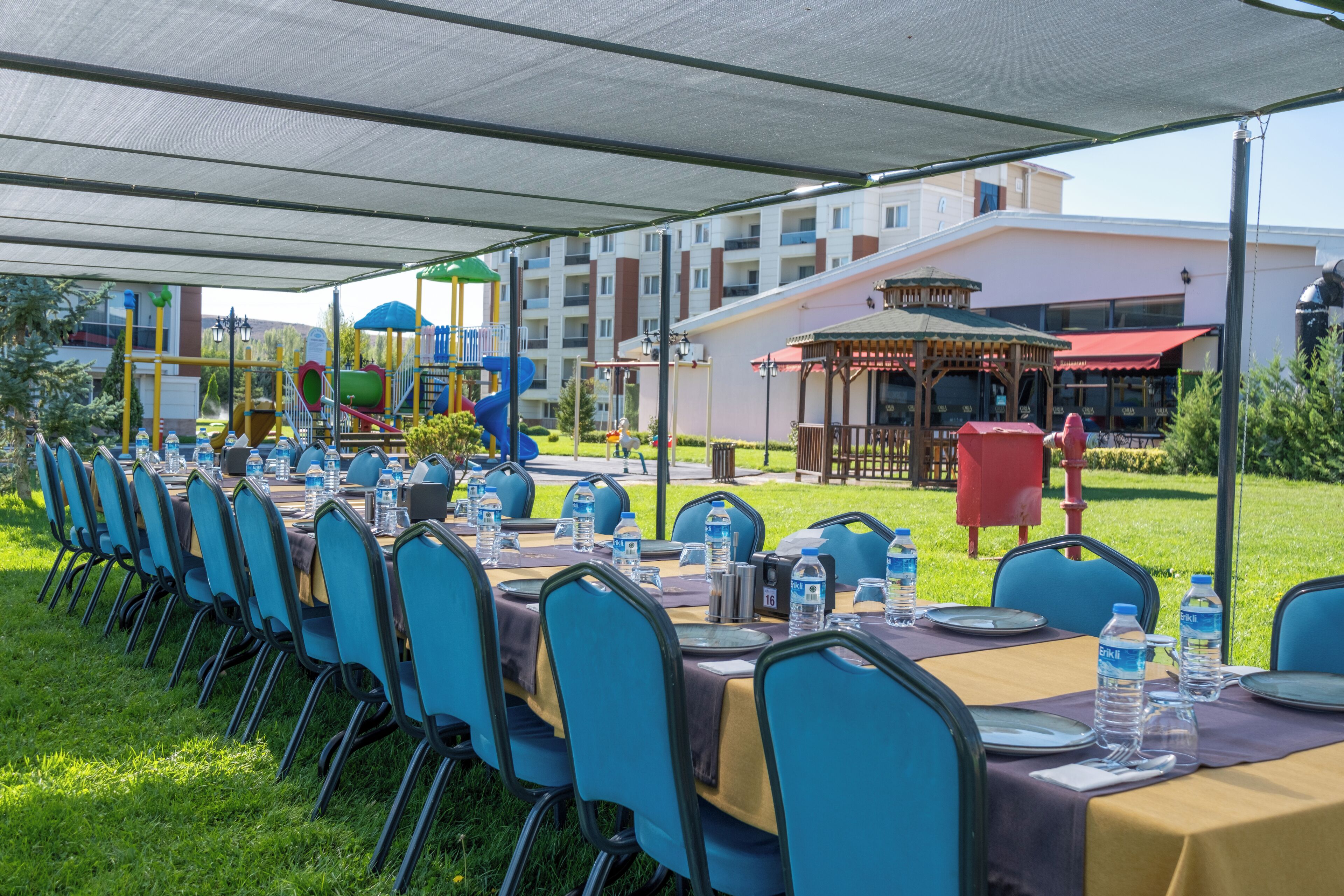 A covered outdoor dining area with tables and chairs, a playground in the background, and a building with balconies.